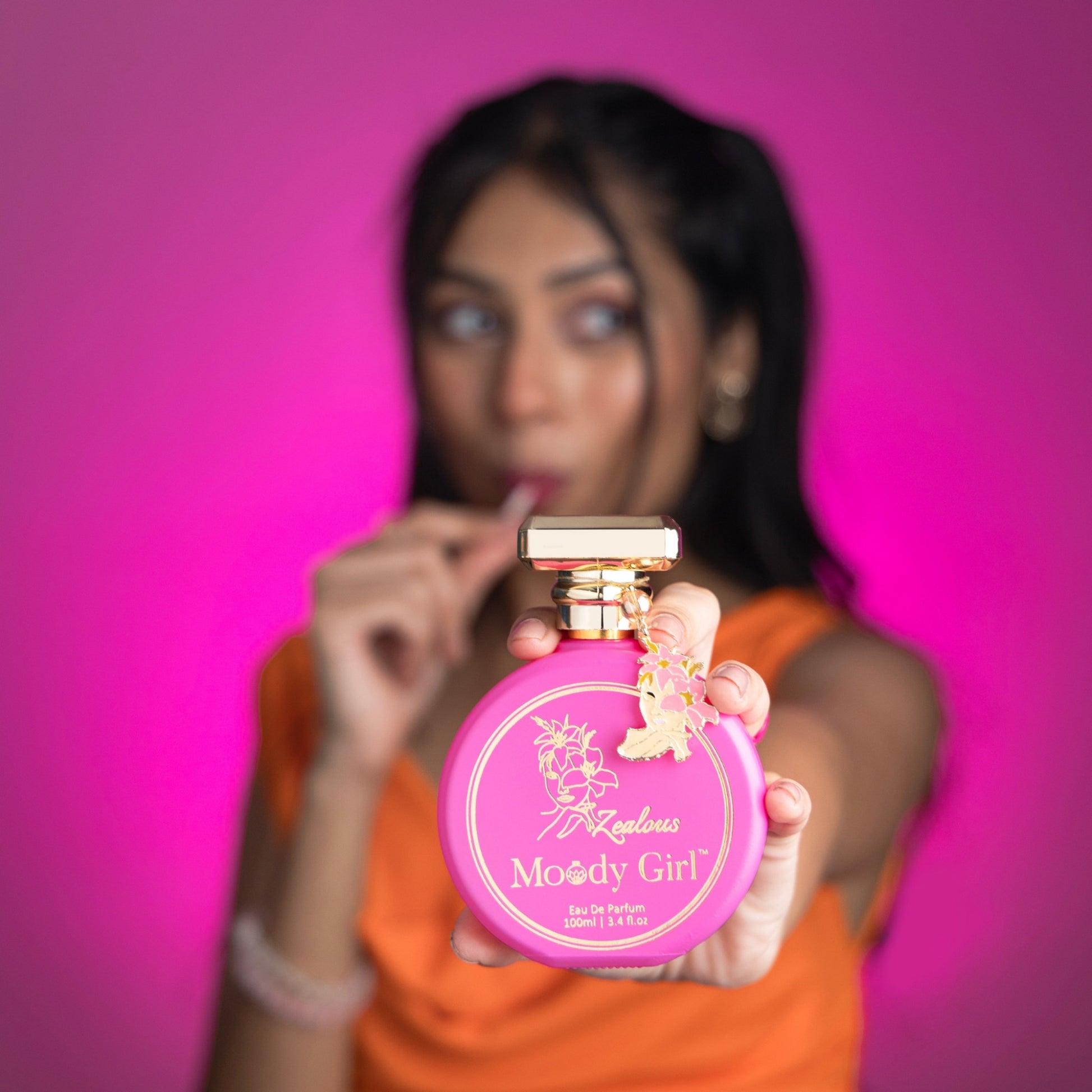 Person holding a Zealous 100ml pink and gold 'Moody Girl' perfume bottle against a pink background
